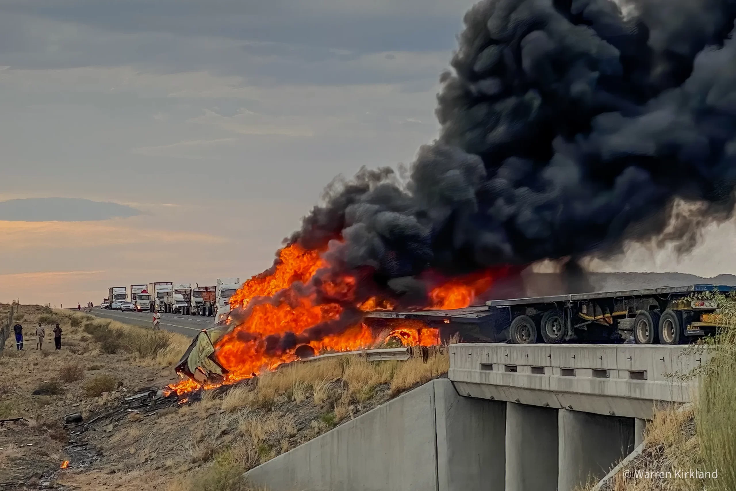 Truck engulfed in flames on a highway overpass, thick black smoke rising