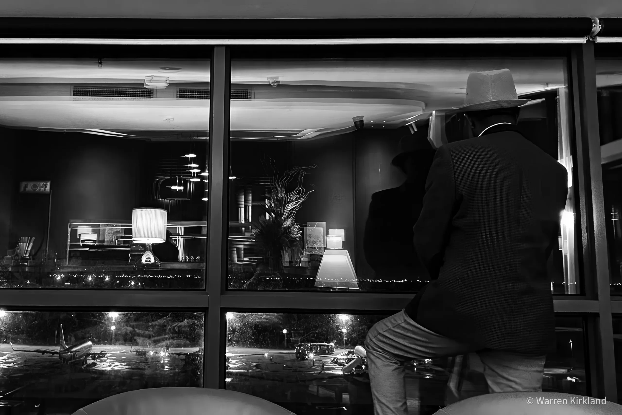 Man in a hat looking out an airport window at night, black and white
