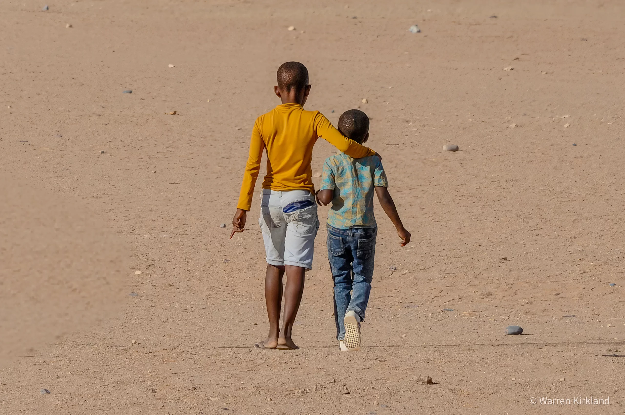 Two children walking together across a sandy landscape, seen from behind