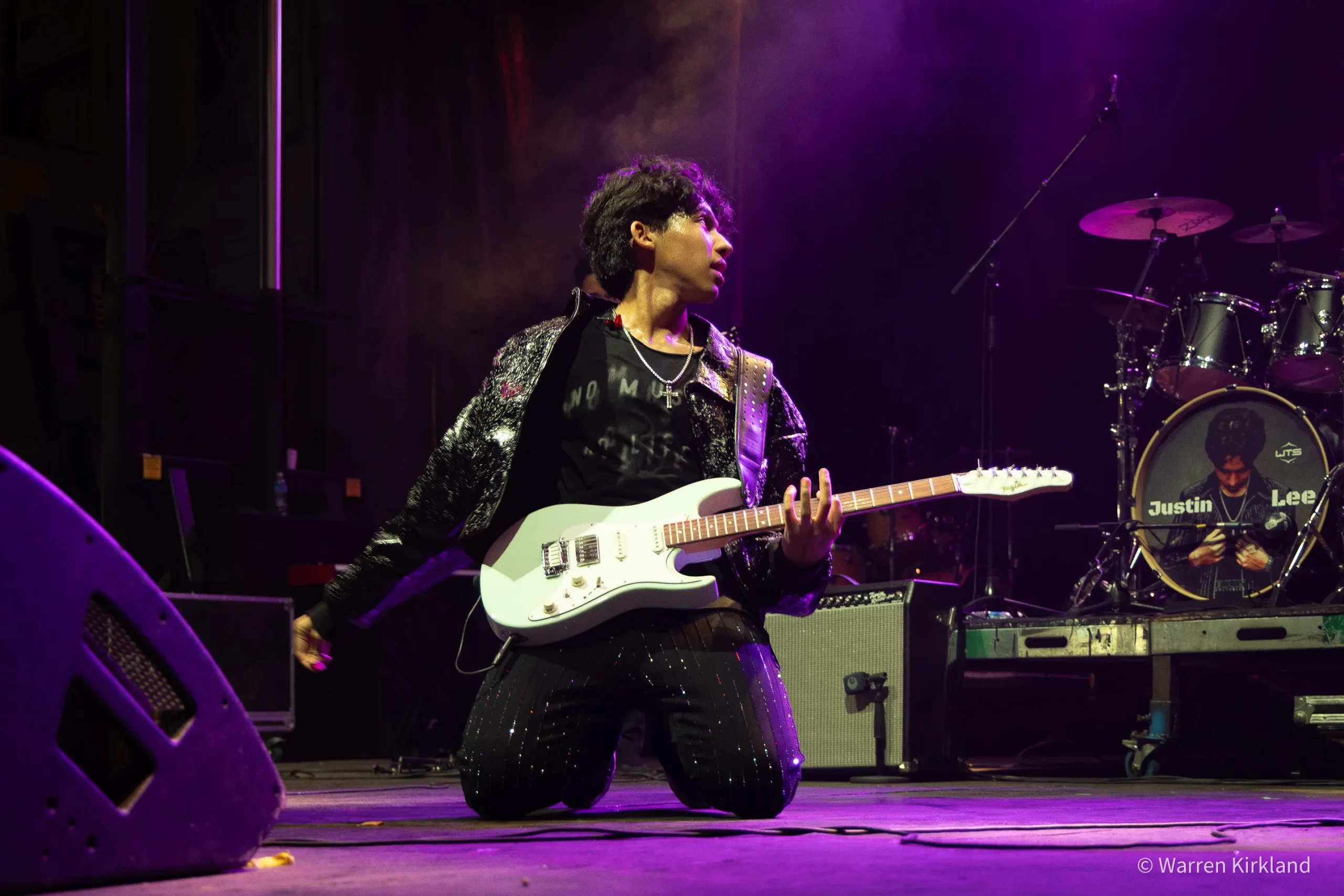 Justin Lee Schultz kneeling on stage playing guitar under purple lights