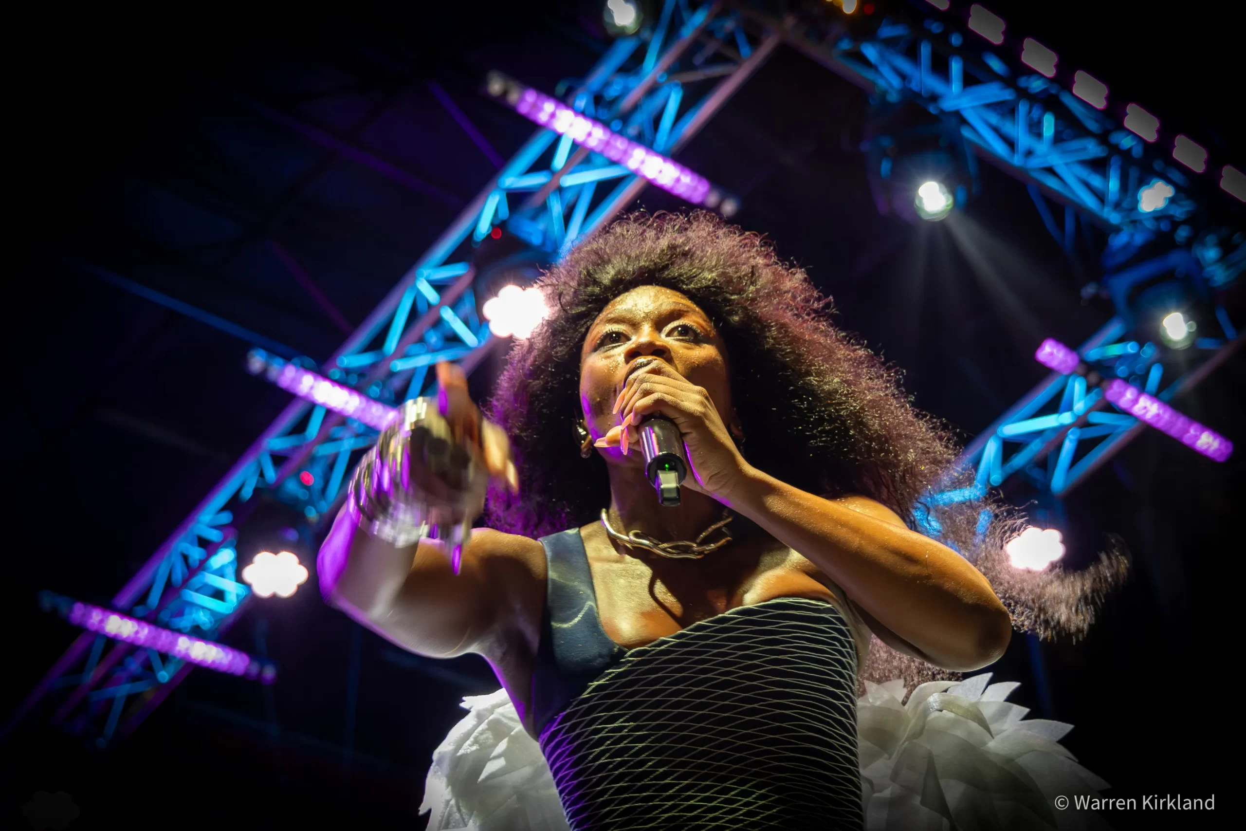Zoë Modiga singing into a microphone against a lit stage truss backdrop