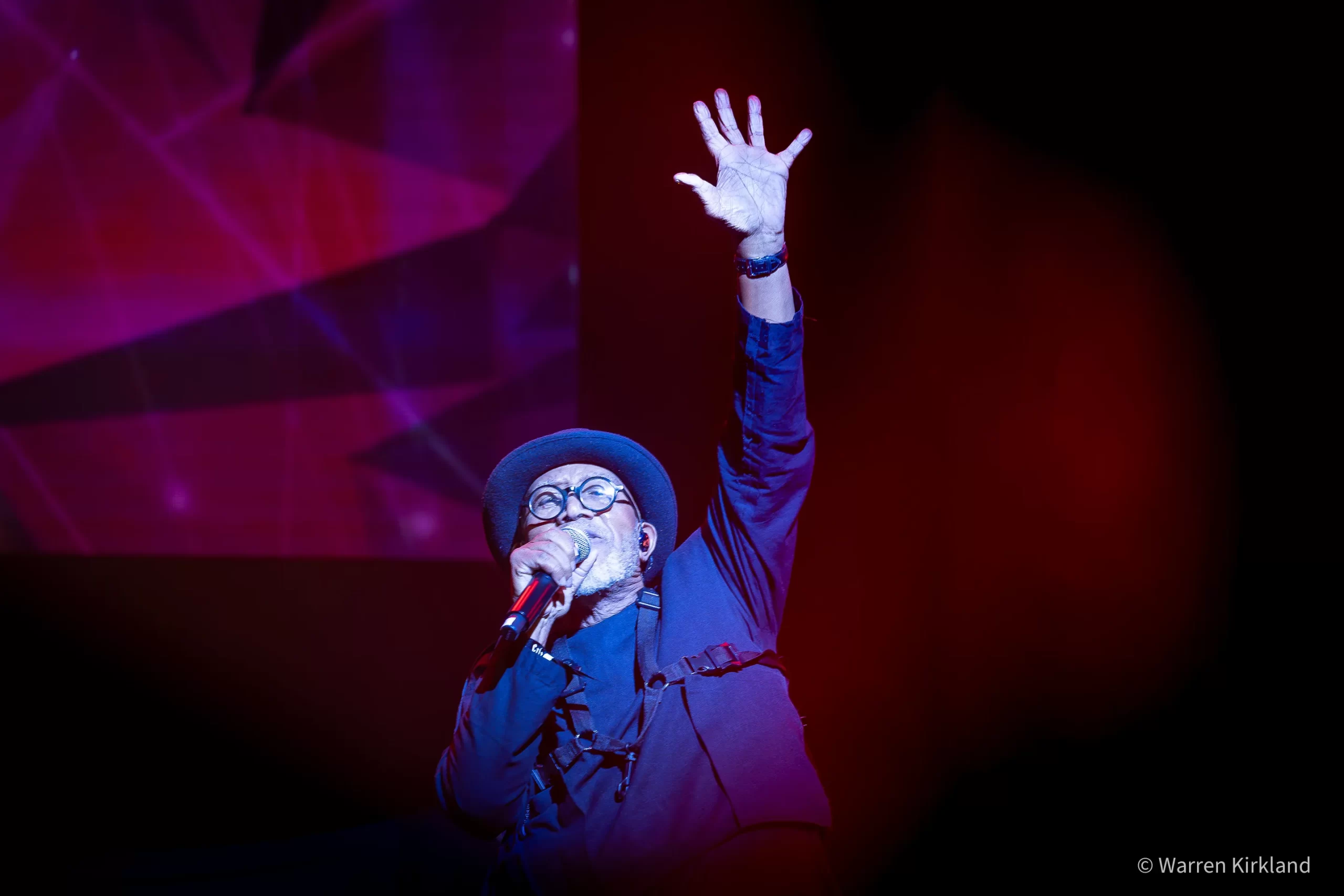 Sipho 'Hotstix' Mabuse singing with hand raised against a dark red backdrop