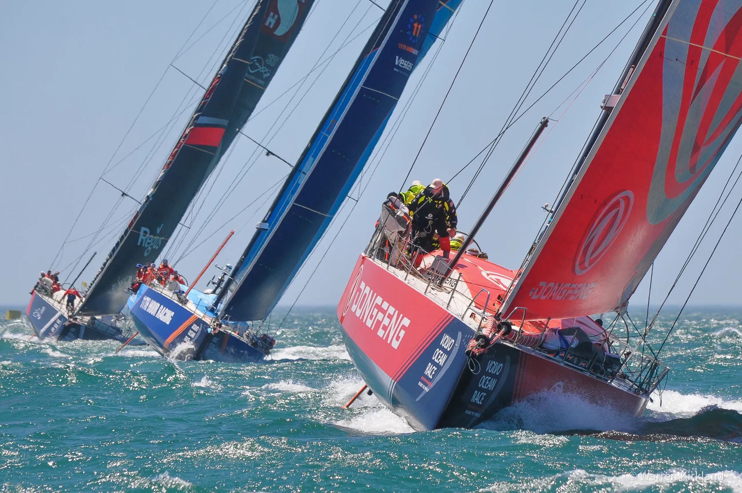 Multiple Volvo Ocean Race yachts sailing close together with spray flying