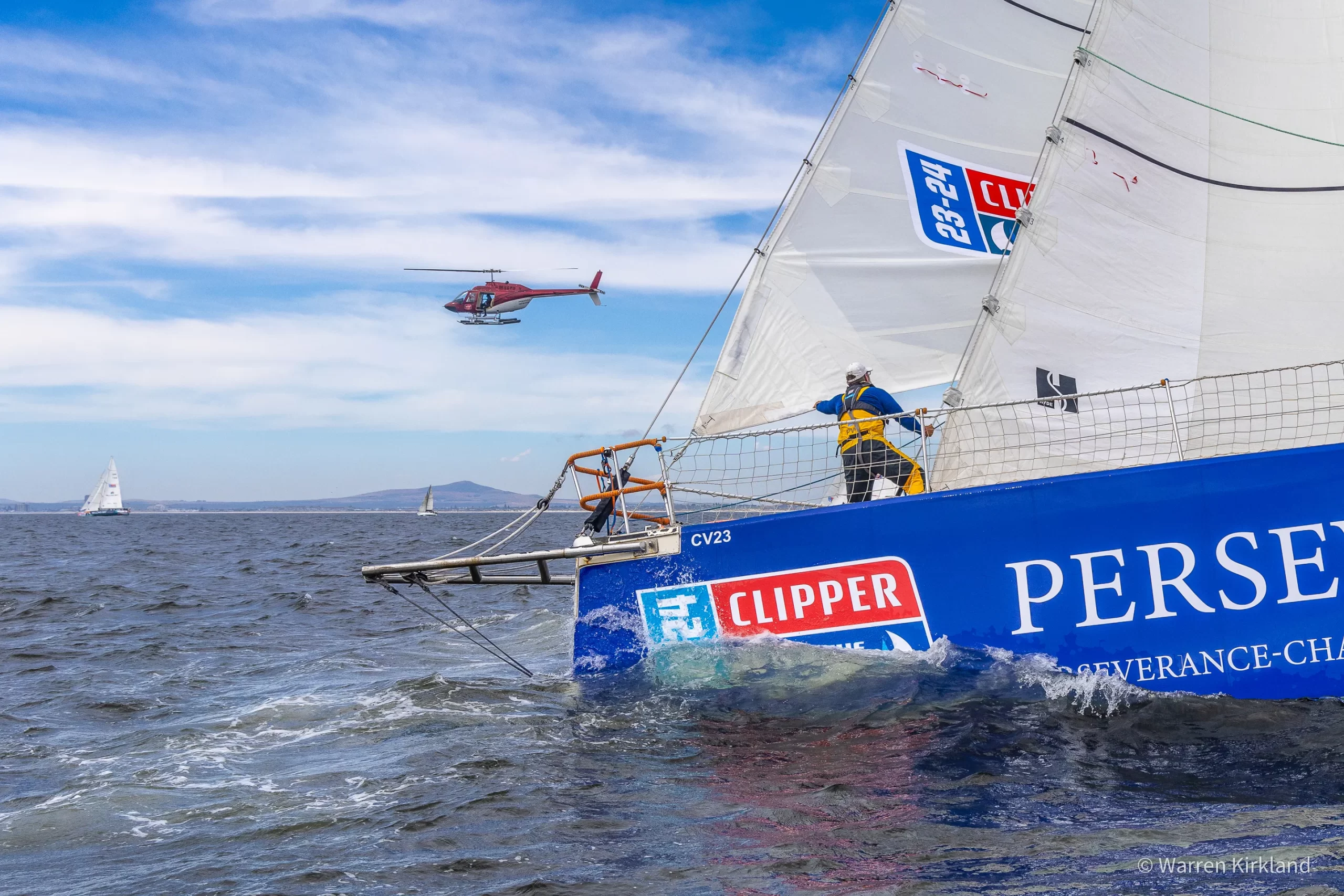 Clipper yacht Perseverance heeling under sail with a helicopter overhead