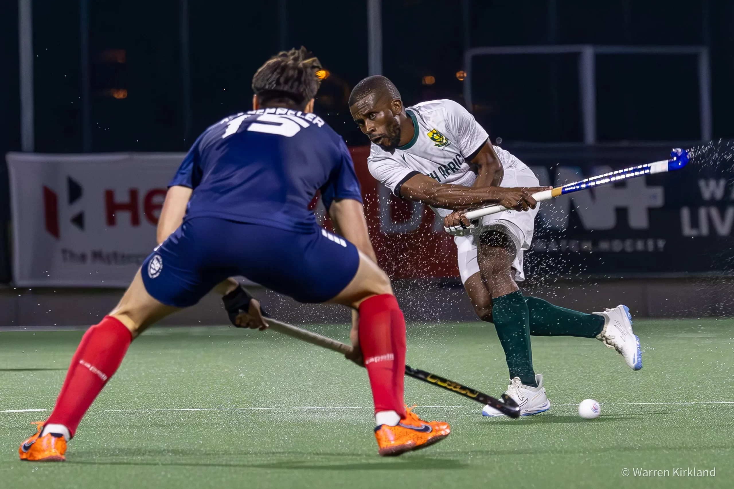 South Africa hockey player dribbling past a defender, water spray from the pitch
