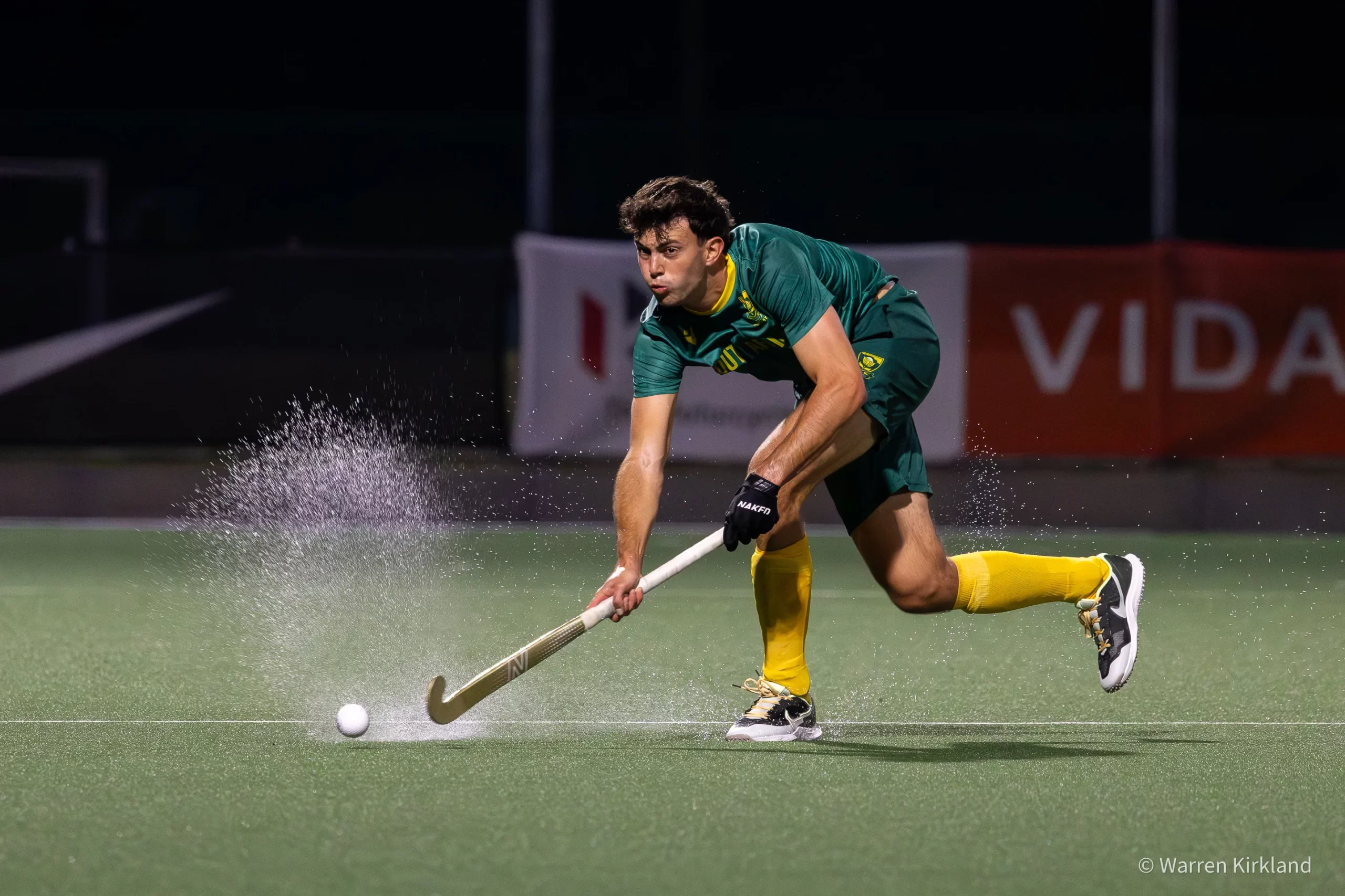 South Africa hockey player striking the ball at speed, water spray behind him